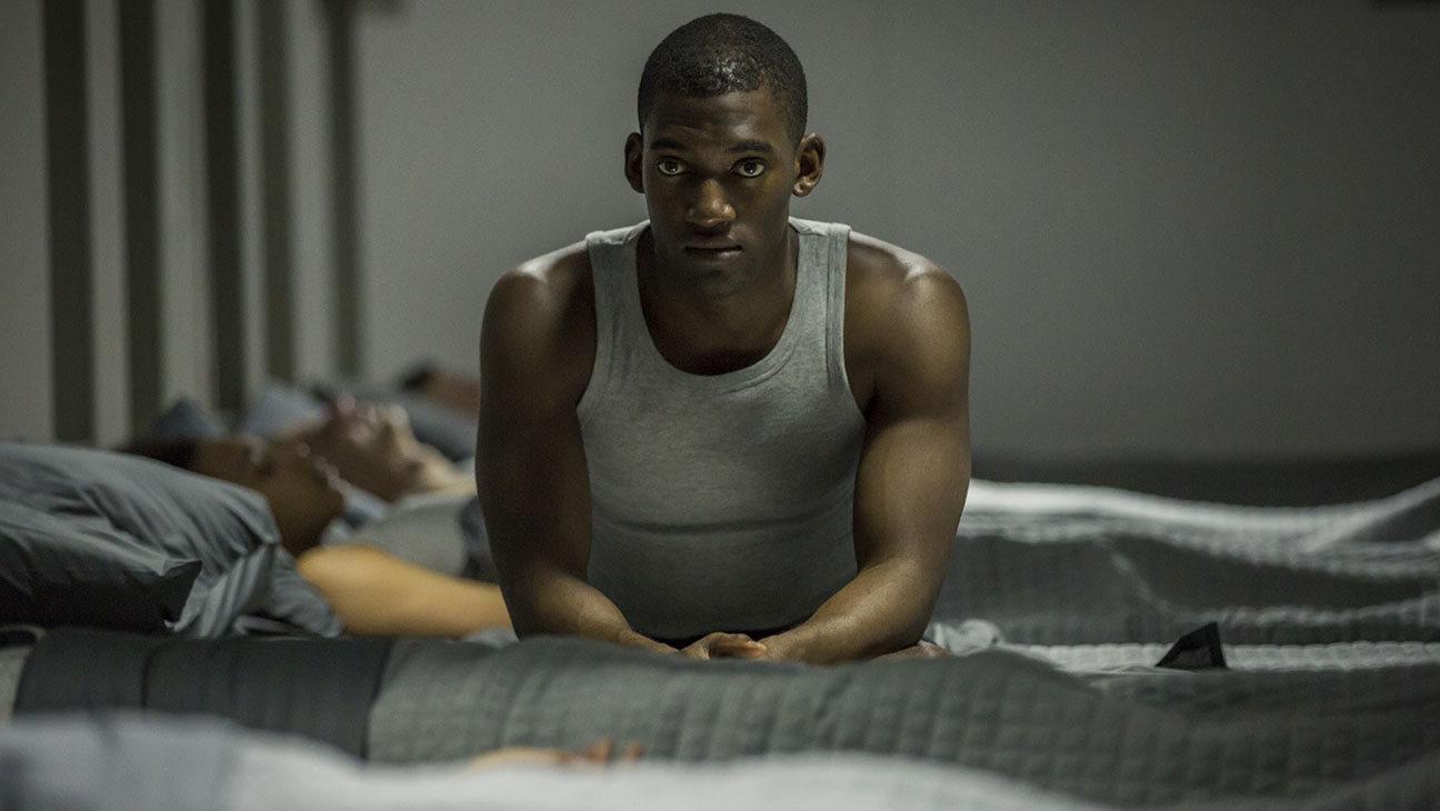 Malachi Kirby, as Stripe in 'Black Mirror', is seated on the edge of his bed, his gaze intense and distant, reflecting deep contemplation or inner turmoil.