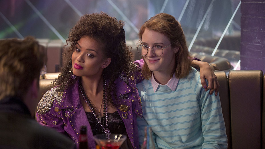 Kelly Booth confidently has her arm around Yorkie, who smiles awkwardly, as they sit together in a booth at a club, capturing a moment of budding connection and contrasting personalities in 'Black Mirror: San Junipero'.
